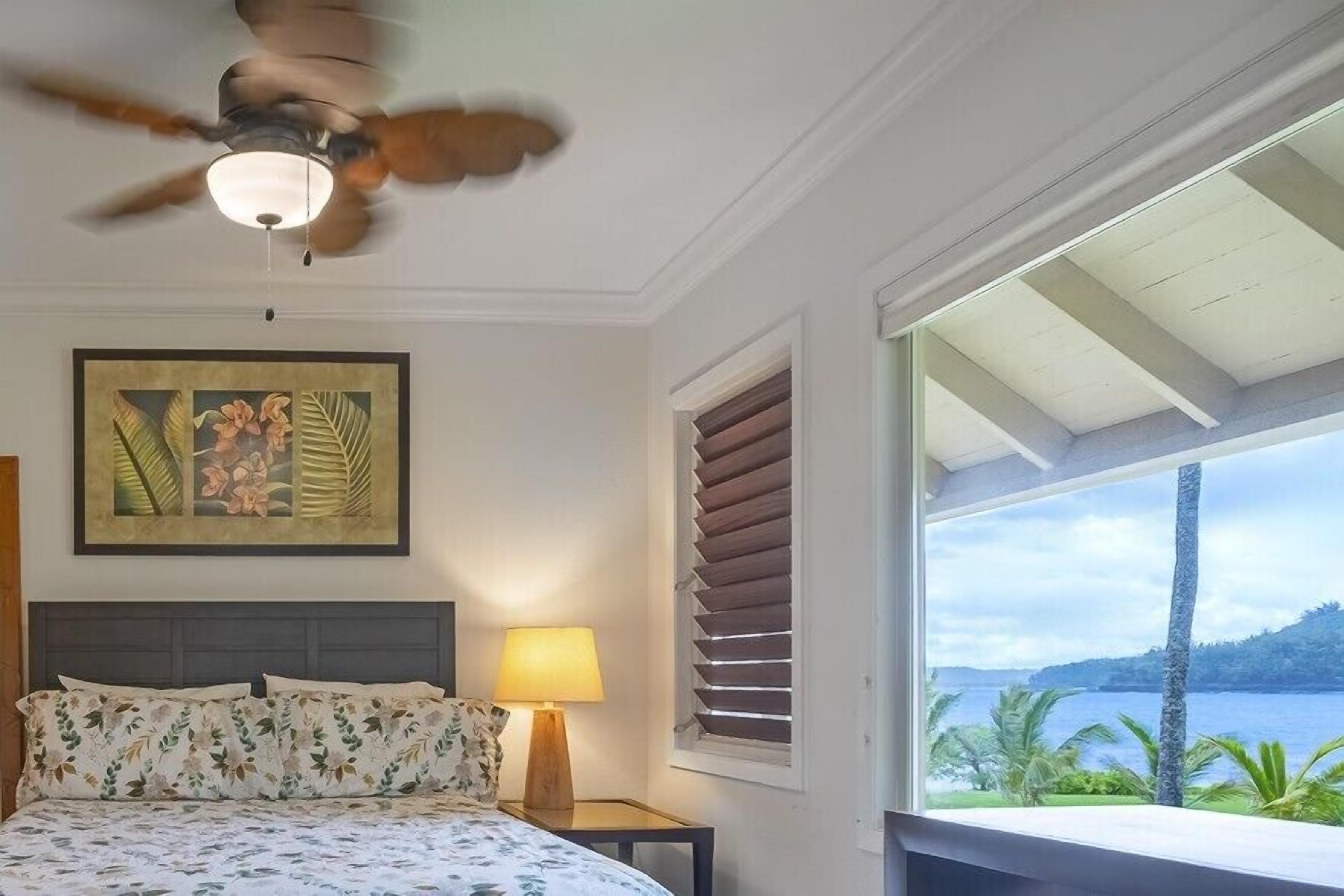 A bedroom with a bed, a ceiling fan, a nightstand, a lamp, a window with blinds, and a view of the outdoors.