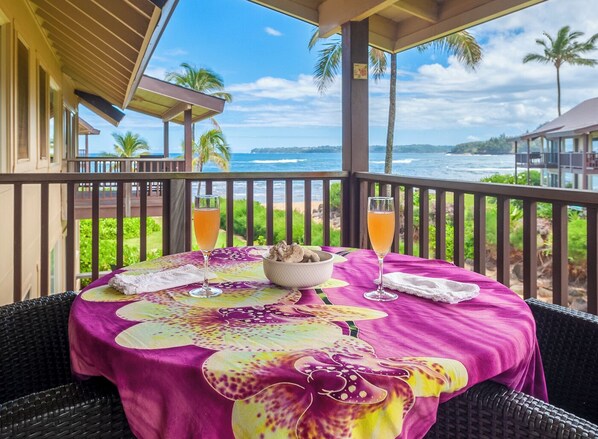 A table with a tropical tablecloth, two drinks, and a bowl of food on a balcony overlooking the ocean. - Hanalei Colony Resort H4 (Hanalei)