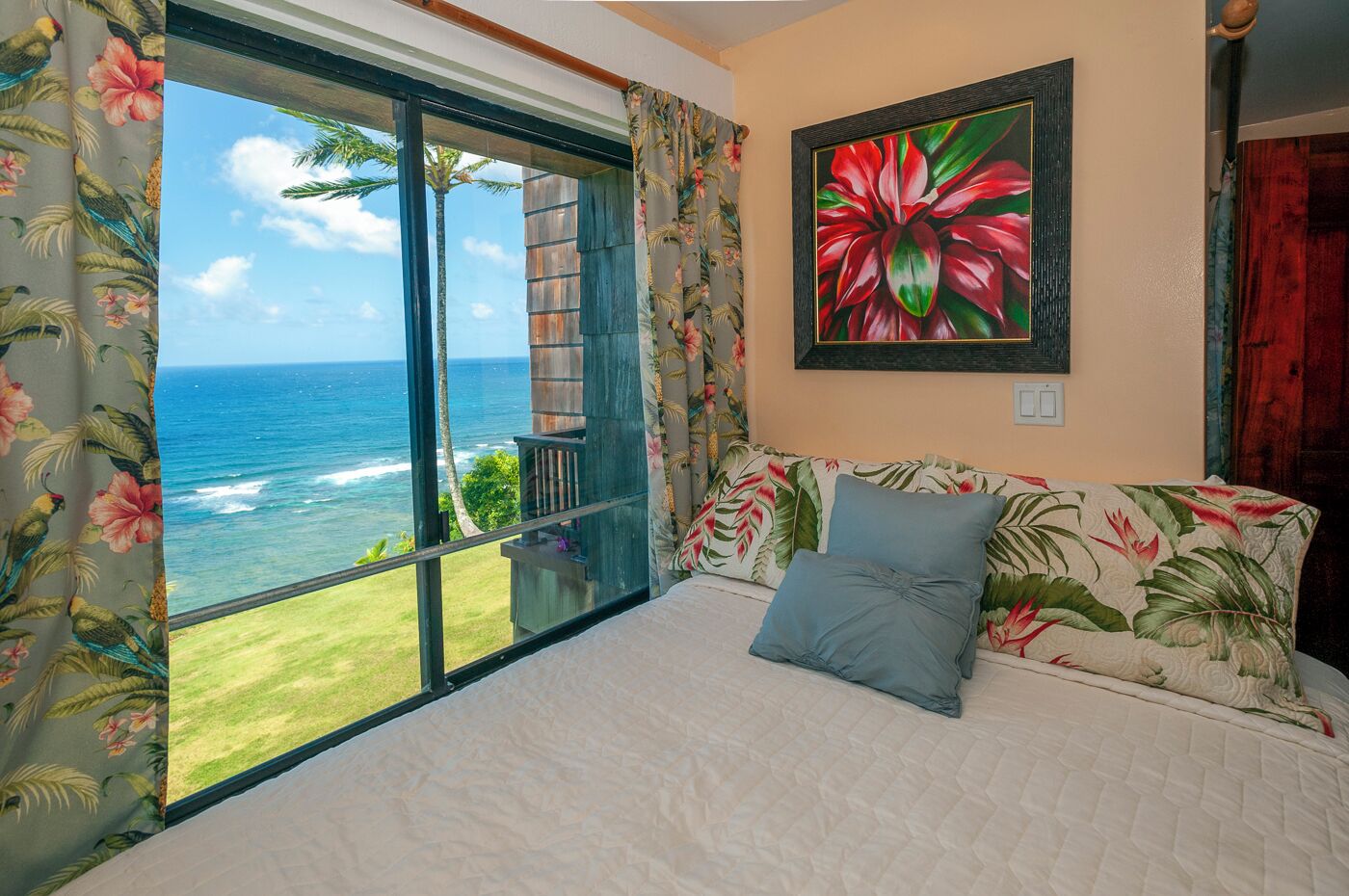 A bedroom with a large window overlooking the ocean, a bed with floral bedding, and a tropical-themed painting on the wall.