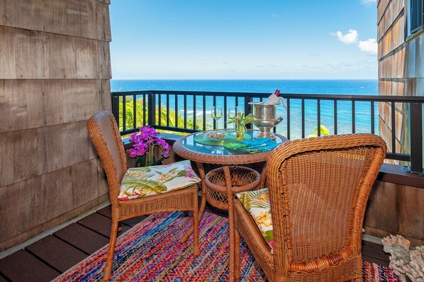 A balcony with wicker chairs, a glass table, and a view of the ocean. - Sealodge A6 (Princeville)