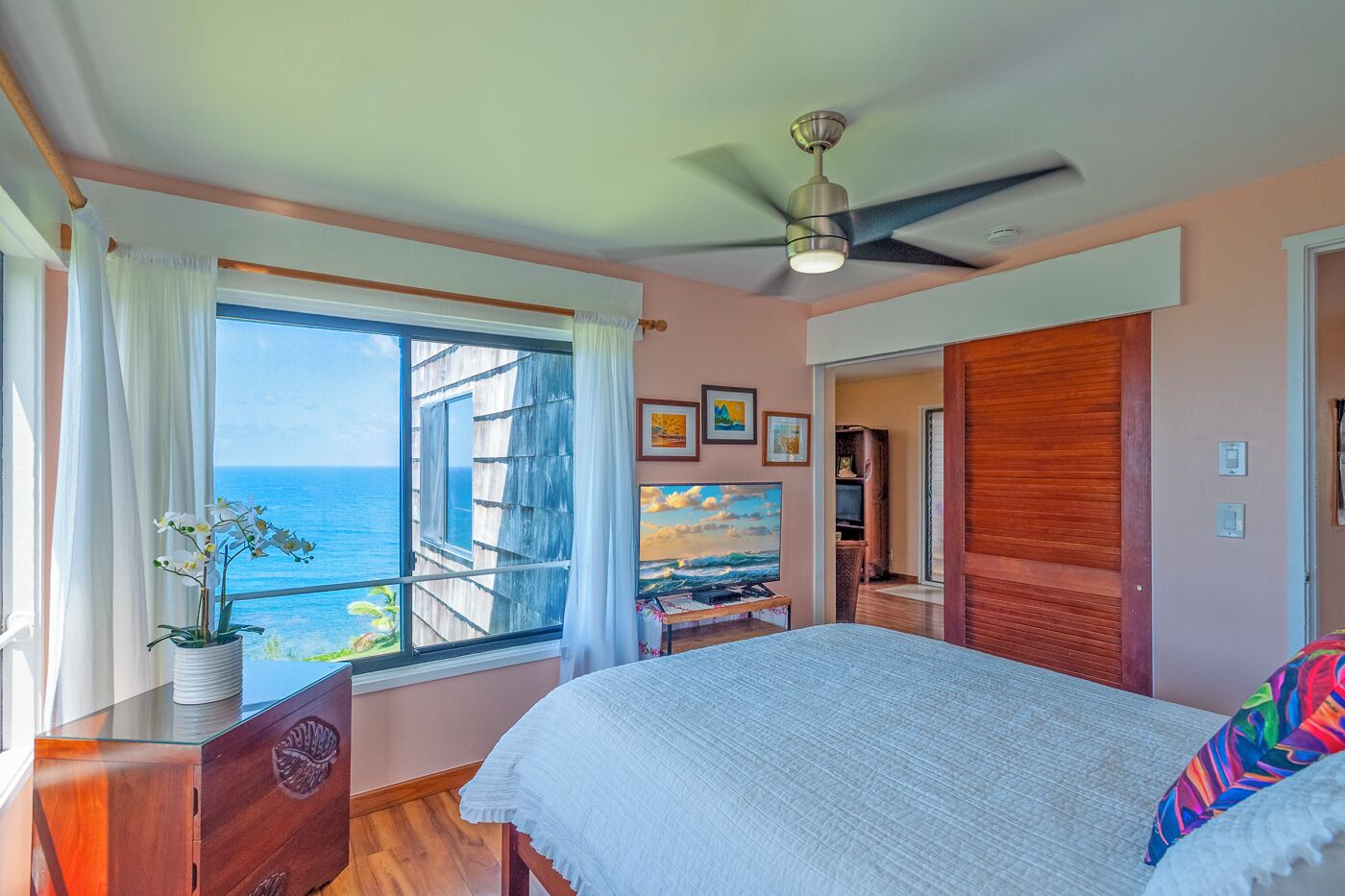 A bedroom with a large bed, a ceiling fan, a window with curtains, a wooden dresser, and a view of the ocean.