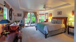 Ein helles Schlafzimmer mit Bett, Nachttischen, Schreibtisch und Stuhl. Ein Fenster mit Vorhängen, ein Deckenventilator und Blick ins Grüne sind ebenfalls vorhanden.