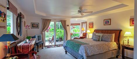 A well-lit bedroom with a bed, bedside tables, a desk, and a chair. There is a window with curtains, a ceiling fan, and a view of greenery outside.