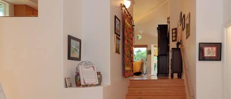 A clean, well-lit hallway with a wooden staircase leading to a room. There are framed pictures and a small table with a vase of flowers on the wall.
