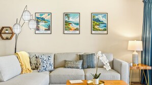 Sala de estar com sofá cinzento, almofadas decorativas, mesa de centro de madeira e três quadros com imagens de praias havaianas.