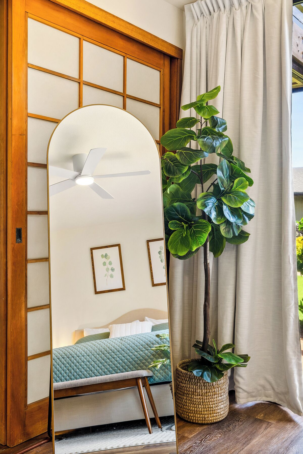 Ein Zimmer mit einem großen Spiegel, der ein Bett, einen Deckenventilator und eine Topfpflanze reflektiert.