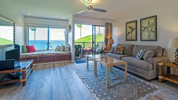 Ein geräumiges Wohnzimmer mit großem Fenster zum Meer, einem Deckenventilator, einer Couch, einem Couchtisch und einem Fernseher.