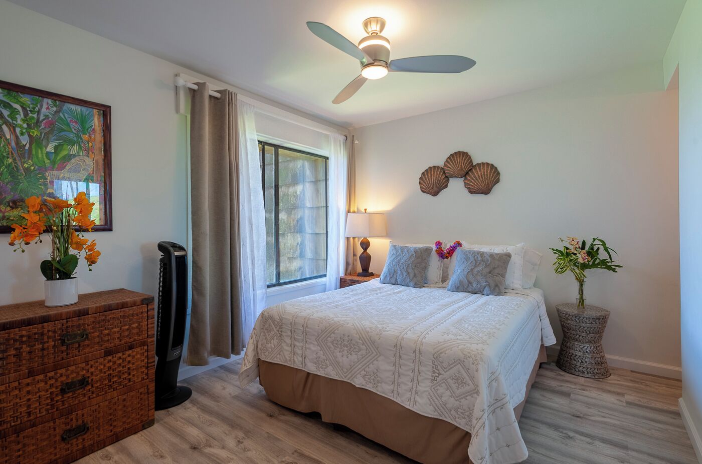 A bedroom with a bed, a ceiling fan, a window with curtains, a bedside table with a lamp, and a wall-mounted decorative piece.
