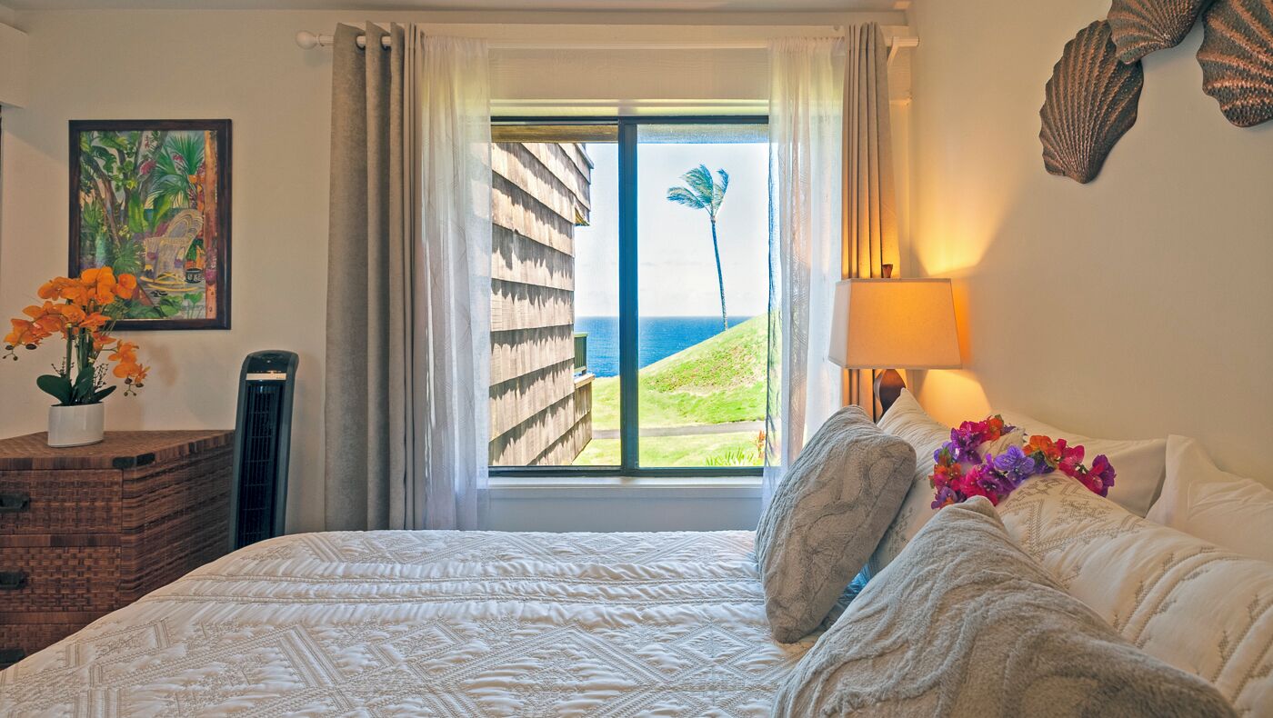 Een slaapkamer met een groot bed, een raam met uitzicht op de oceaan, een kamerplant en een lamp.