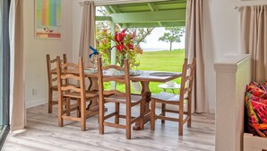A dining area with a wooden table and chairs, a hanging light fixture, and a view of a green lawn and trees through a window. - Kamahana 16 - Downstairs (Princeville)