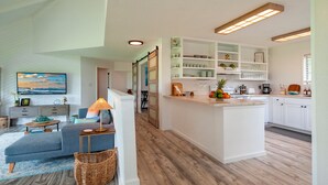 A modern living room with a sofa, coffee table, and a lamp. A kitchen with white cabinets and a central island is visible. - Kamahana 16 - Downstairs (Princeville)