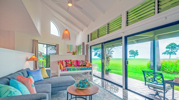 A modern living room with a sofa, a coffee table, and a view of a green landscape through large glass doors. - Kamahana 16 - Downstairs (Princeville)