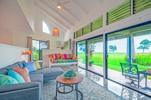 A modern living room with a sofa, a coffee table, and a view of a green landscape through large glass doors. - Kamahana 16 - Downstairs (Princeville)