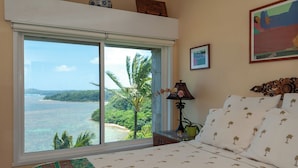 A bedroom with a large window offering a view of the ocean and palm trees. - Sealodge E10 (Princeville)