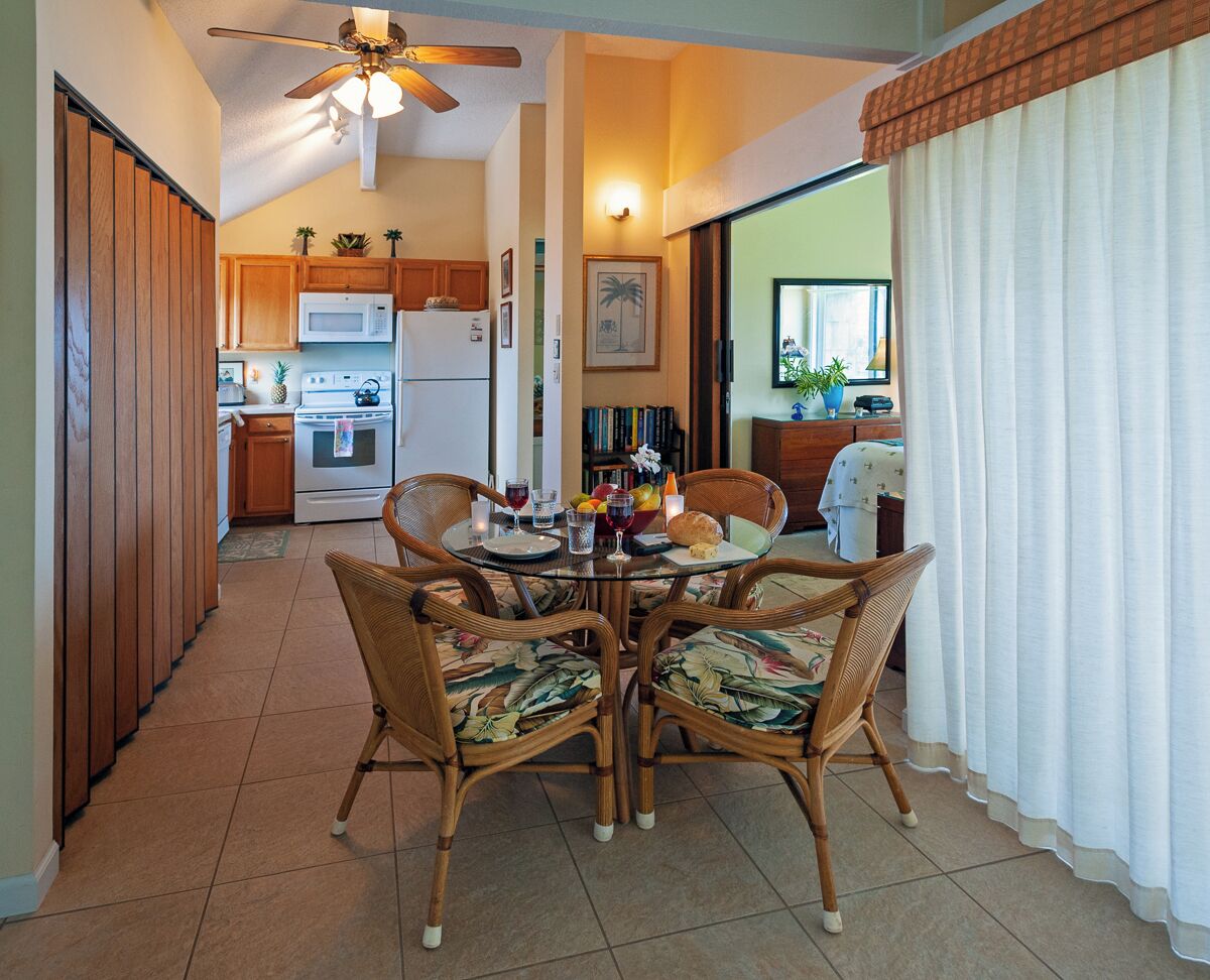 A dining area with a round table and four wicker chairs, a ceiling fan, and a kitchen in the background.