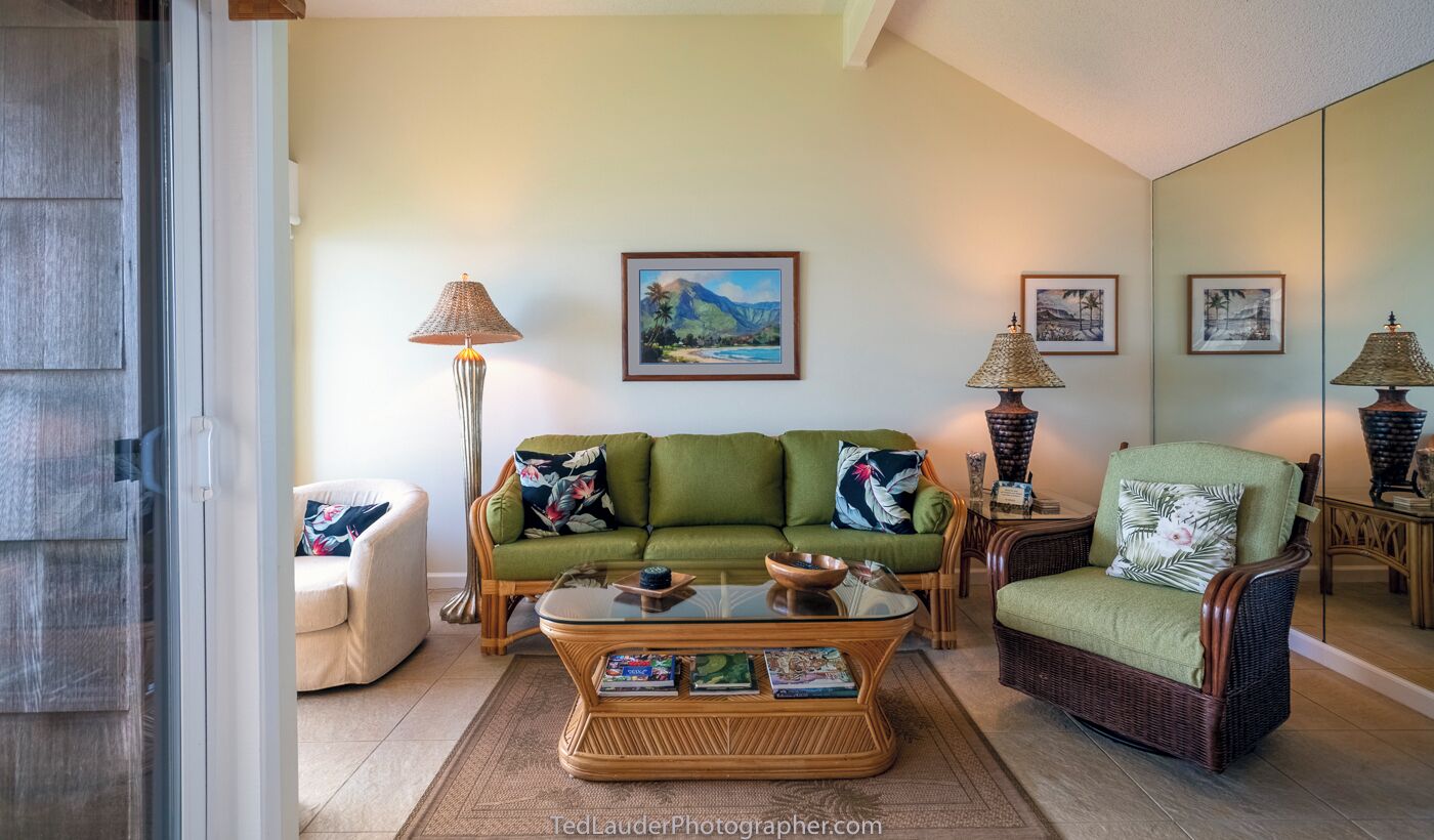 Un salon avec un canapé vert, un fauteuil en osier, une table basse en verre sur laquelle sont posés des livres, et deux lampes fixées au mur.