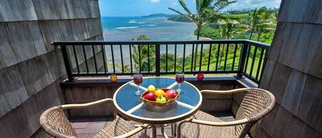 A balcony with wicker chairs, a glass table with a fruit bowl, and a view of the ocean and palm trees.