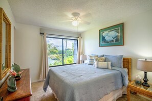 A bedroom with a large bed, a ceiling fan, a window with a view of greenery, a framed picture on the wall, and a bedside table with a lamp.