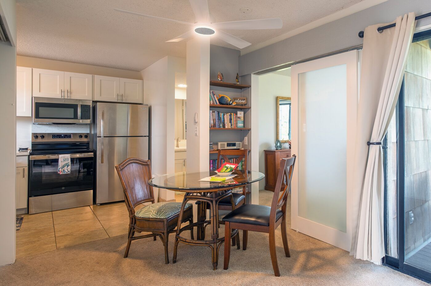 Una cucina compatta con elettrodomestici moderni, una zona pranzo con tavolo in vetro e vista all'esterno tramite una porta scorrevole.