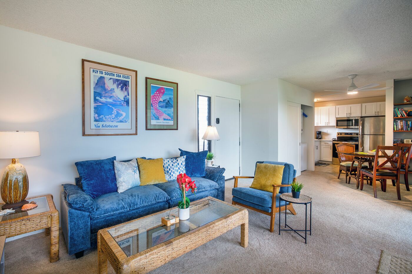 A living room with a blue sofa, a coffee table, and a dining area with chairs and a table. There are framed pictures on the wall, a lamp, and a bookshelf.