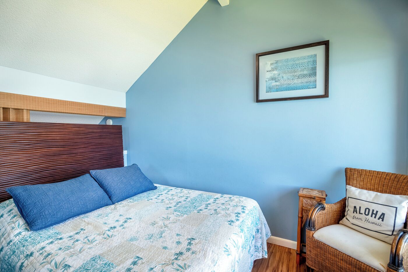 A bedroom with a bed, a chair, a framed picture, and a pillow with 'ALOHA from Hawaii'.