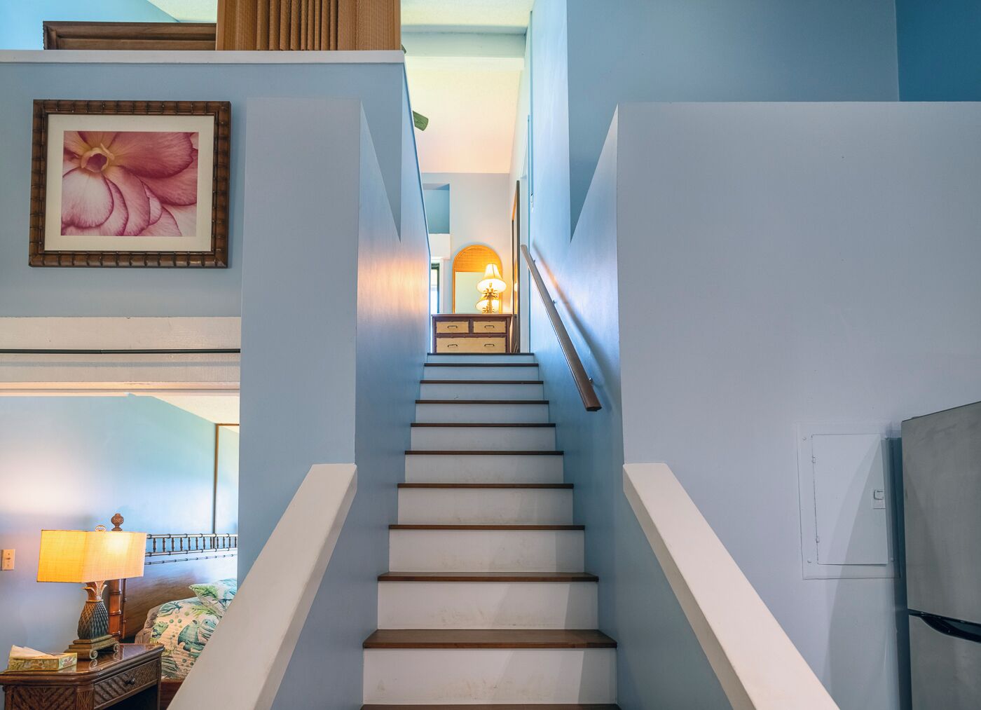 Una escalera con peldaños blancos y barandillas de madera que conduce a un rellano con una lámpara y un espejo.