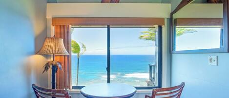 A room with a view of the ocean, a table with chairs, a lamp, and a small shelf with decorative items.