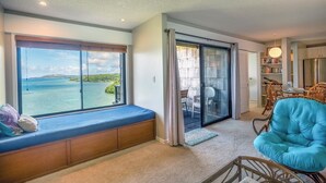 A room with a large window overlooking a body of water, a sliding glass door leading to an outdoor area, and a seating area with a blue chair and wooden bench. - Sealodge E8 (Princeville)