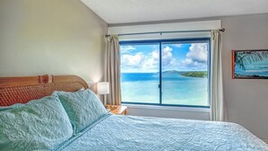 A hotel room with a bed, a nightstand, a window with a view of the ocean, and a painting on the wall. - Sealodge E8 (Princeville)