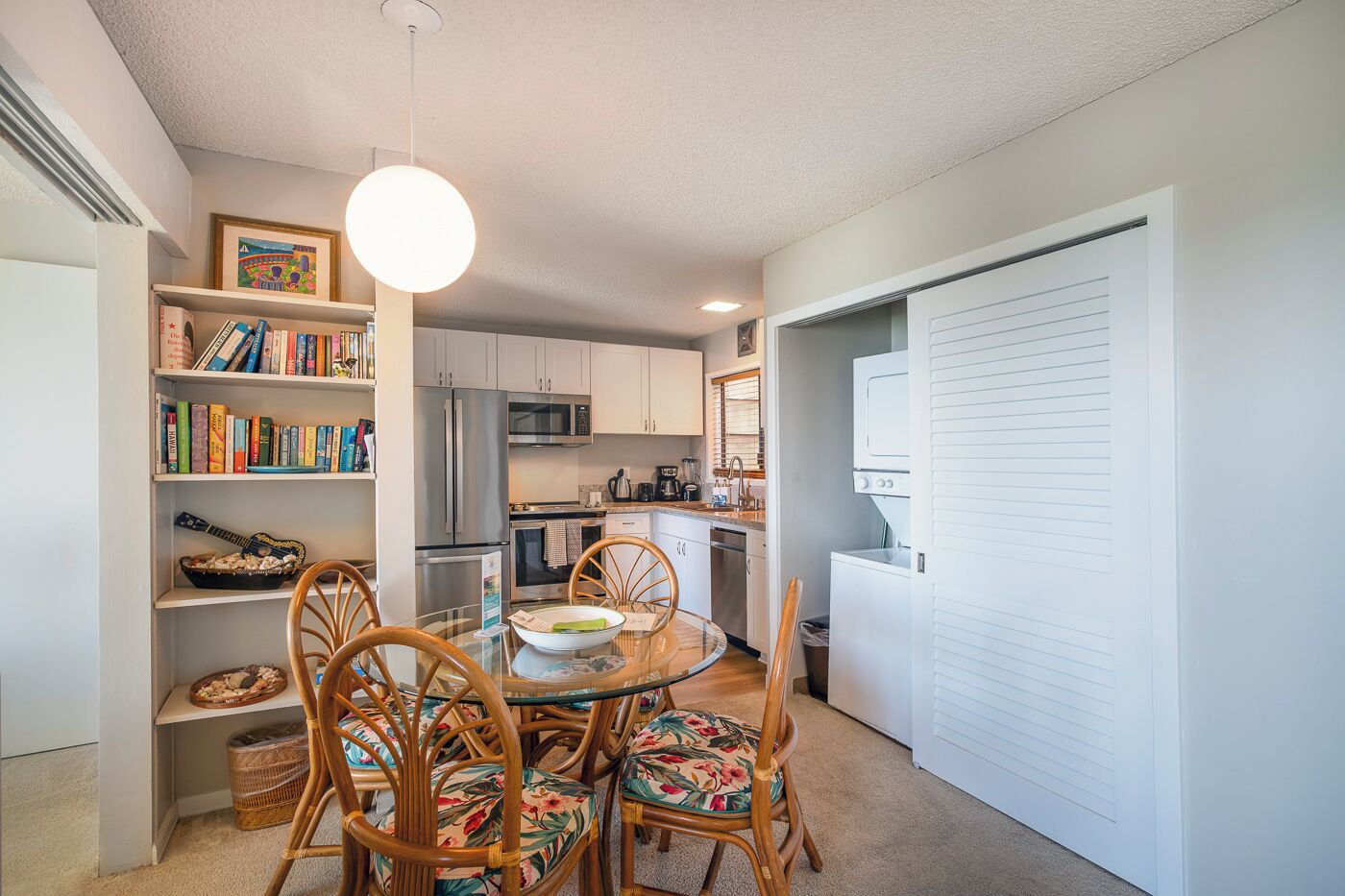 A compact kitchen with modern appliances, a dining area with a glass table, and a bookshelf filled with books.