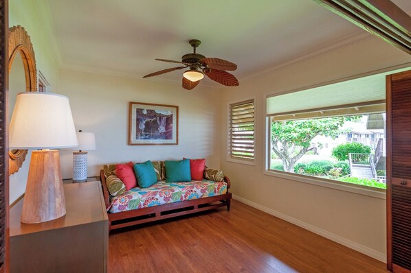 A room with a wooden floor, a ceiling fan, a sofa with colorful cushions, a lamp, a framed picture on the wall, and a window with blinds. - Hanalei Colony Resort K4 (Hanalei)