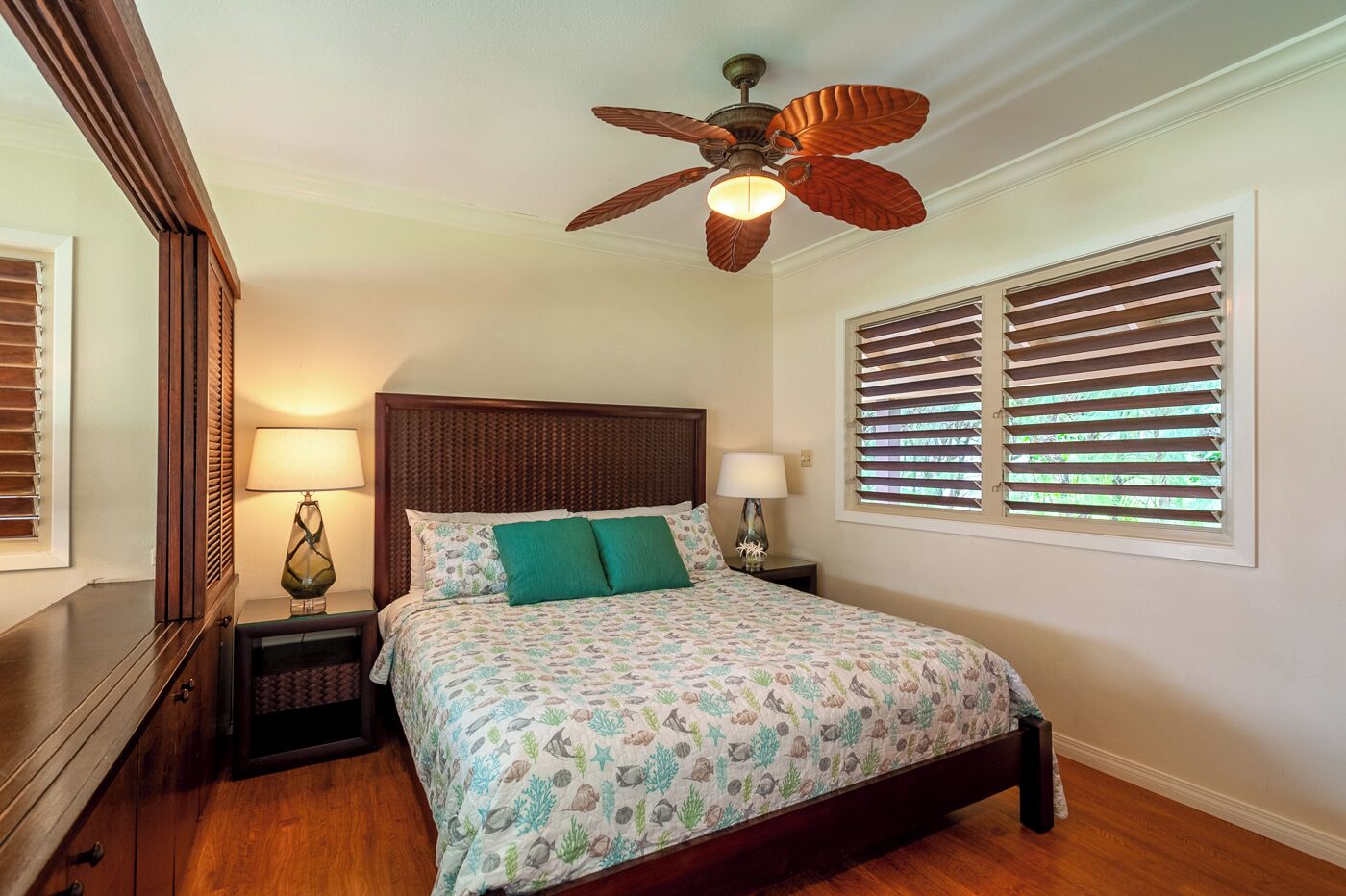 Une chambre à coucher avec une tête de lit en bois, deux tables de chevet avec des lampes, un ventilateur de plafond et une fenêtre avec des stores.