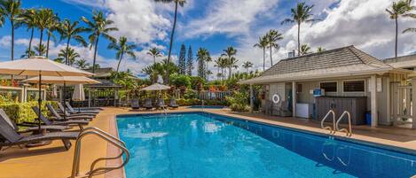 A swimming pool with lounge chairs and umbrellas, surrounded by palm trees and a building with a tiled roof.