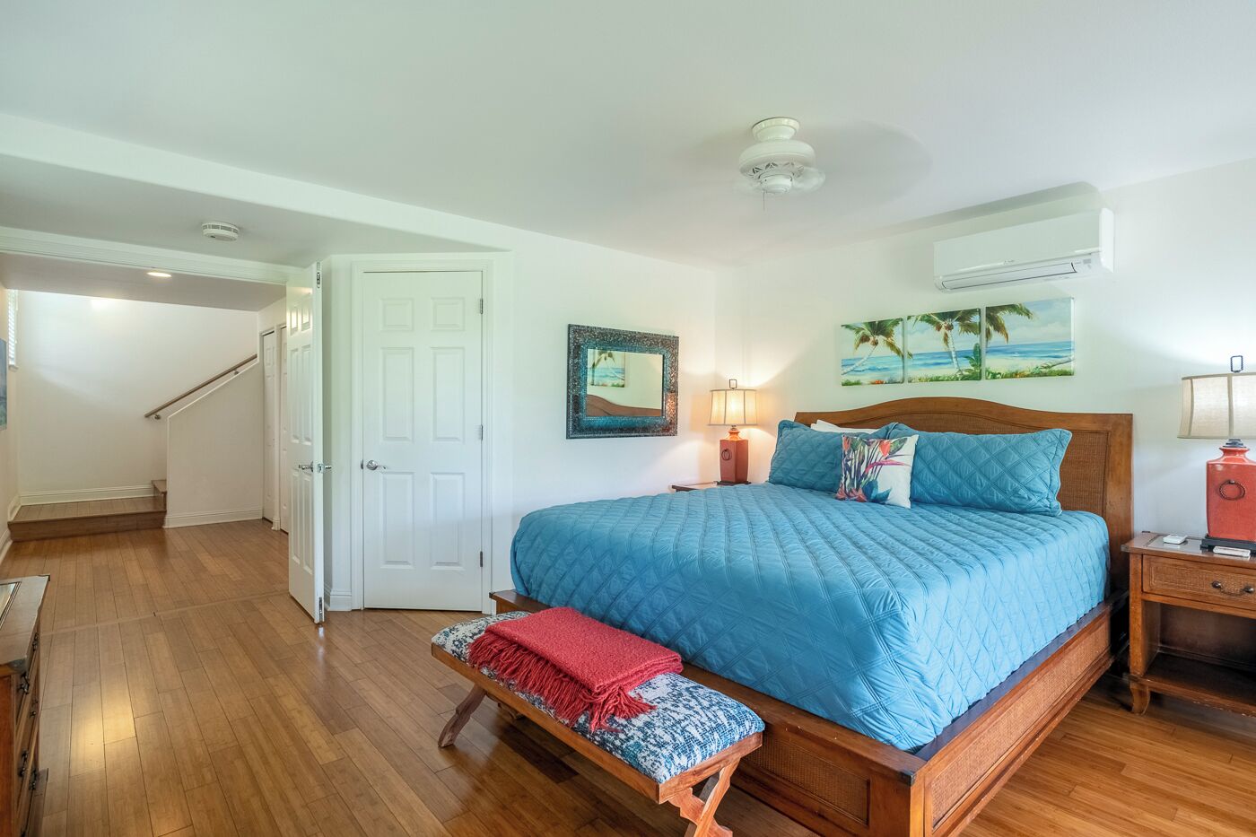 A bedroom with a large bed, a wooden bench, a nightstand, a wall-mounted air conditioner, and a door leading to another room.