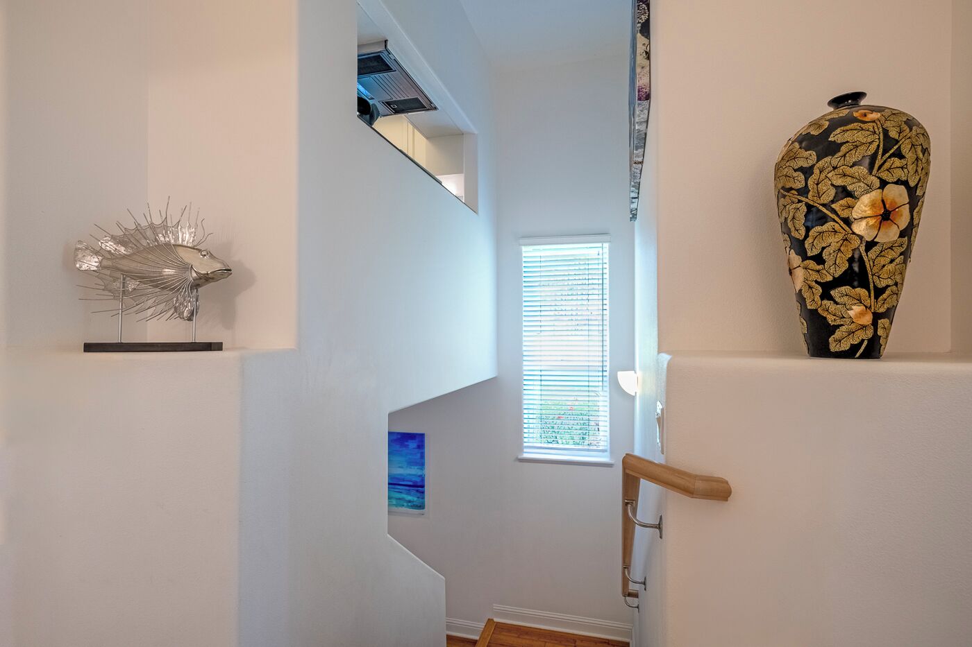 Un corridoio con una moderna scala, una scultura a forma di pesce decorativa, un vaso con motivi floreali e una finestra con persiane.