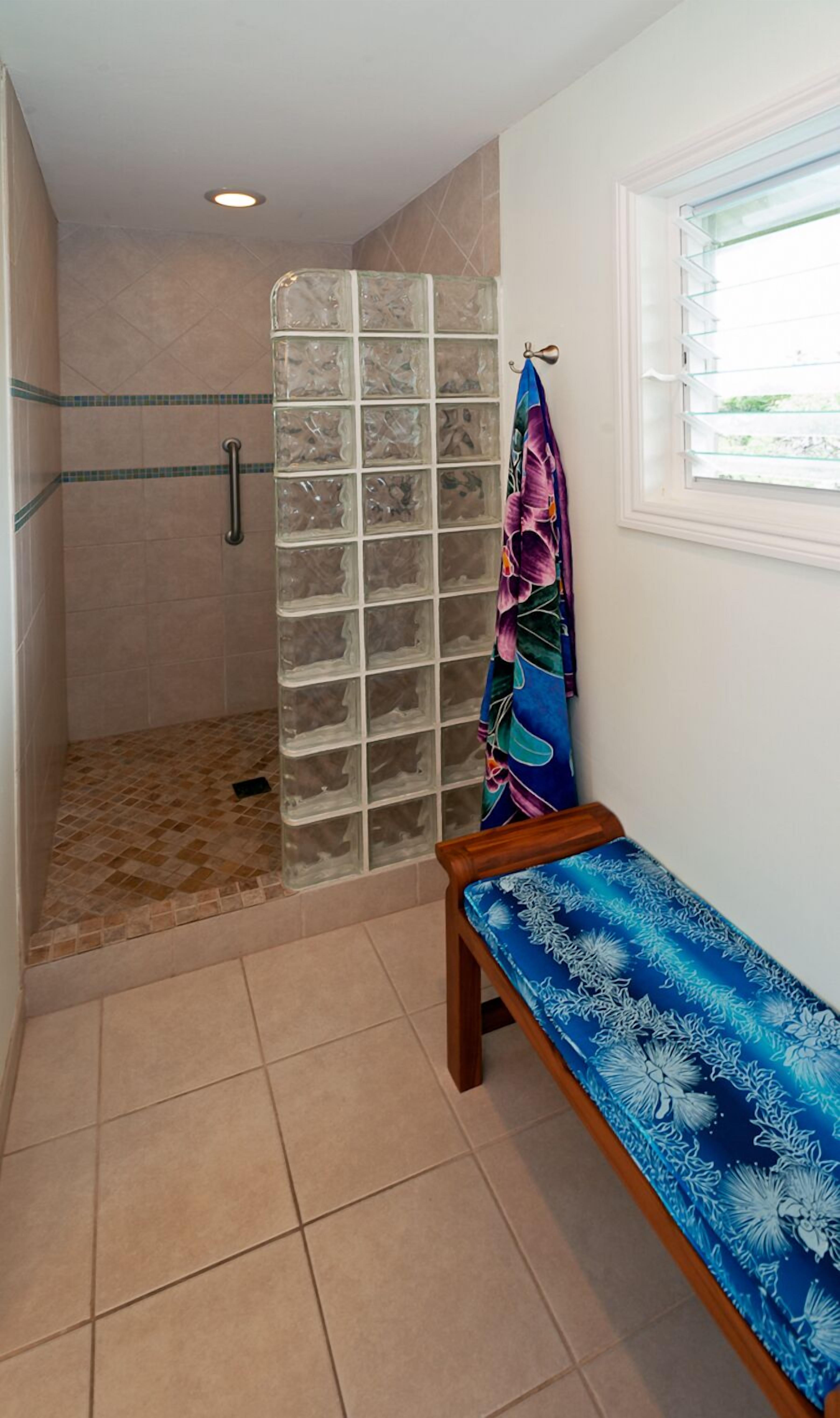 A bathroom with a shower area, a bench, and a window with blinds.
