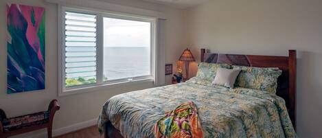 A bedroom with a bed, a ceiling fan, a window with blinds, a nightstand with a lamp, and a chair.