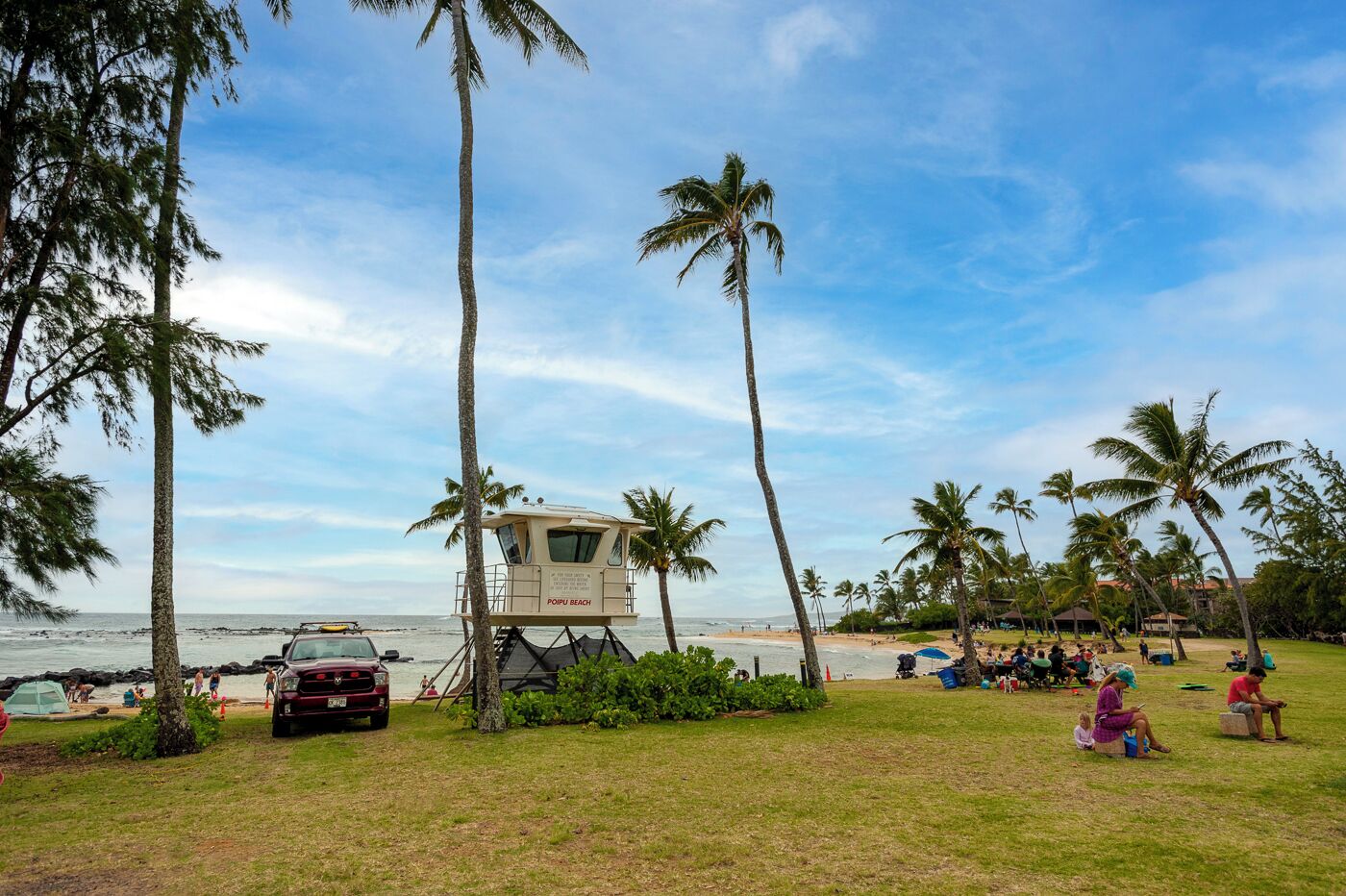 A lifeguard tower at Poipu Beach with a red vehicle parked nearby, surrounded by palm trees and beachgoers.