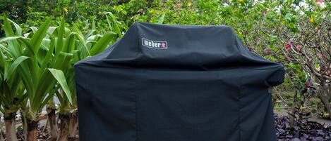 A portable Weber grill covered with a black cloth, placed outdoors with lush greenery and flowering plants in the background.