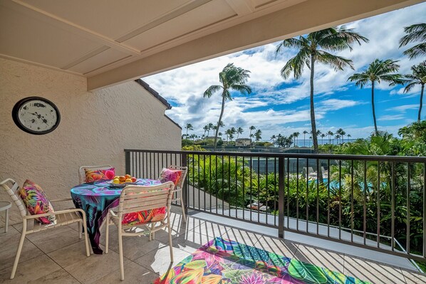 A balcony with a table and chairs, a clock on the wall, and a view of palm trees and a pool. - Kahala at Poipu Kai 123 (Koloa)