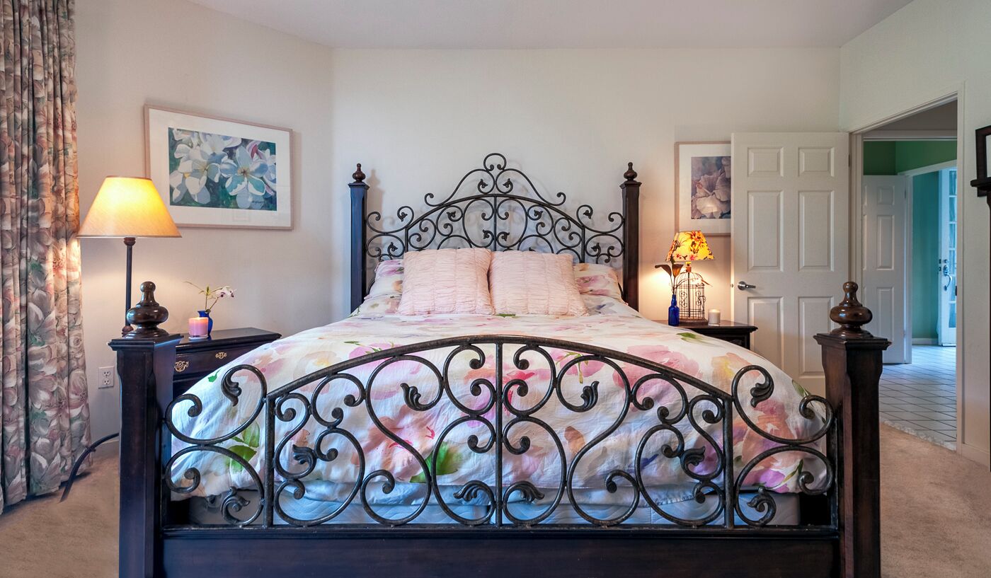 Uma cama bem arrumada com colcha florida, cabeceira de metal decorativa e duas luminárias de apoio.