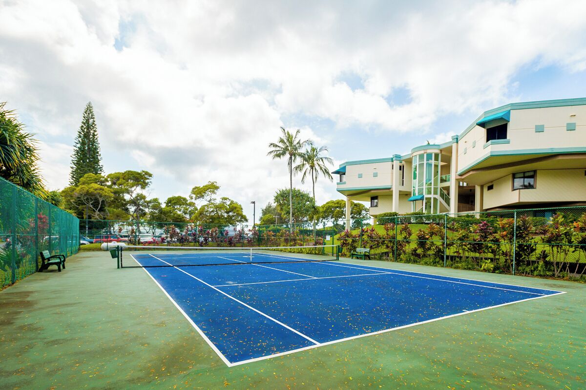 A tennis court with a net, surrounded by a fence, and adjacent to a building with balconies.
