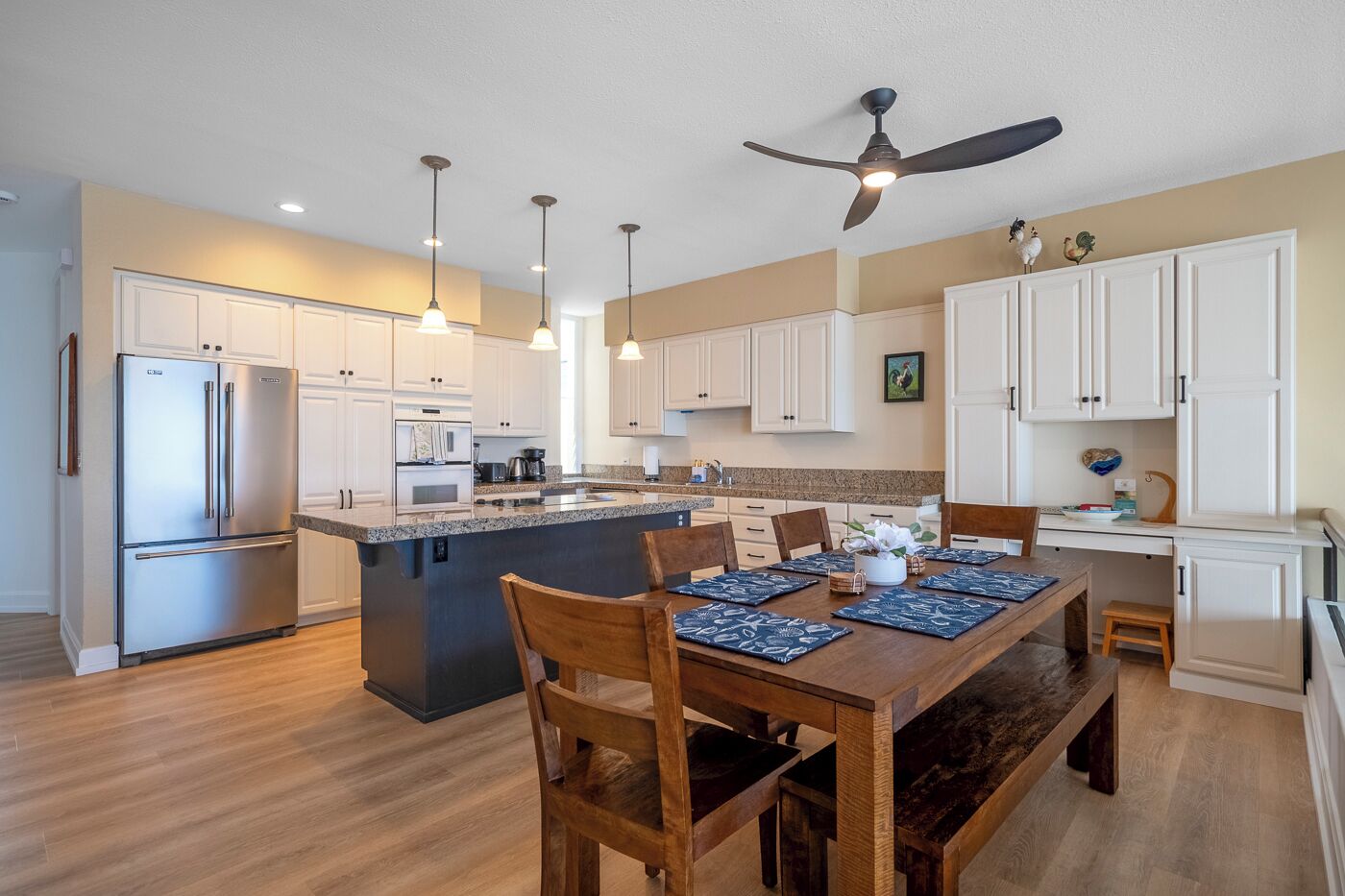 A modern kitchen with white cabinets, a stainless steel refrigerator, a central island with seating, pendant lights, and a ceiling fan.