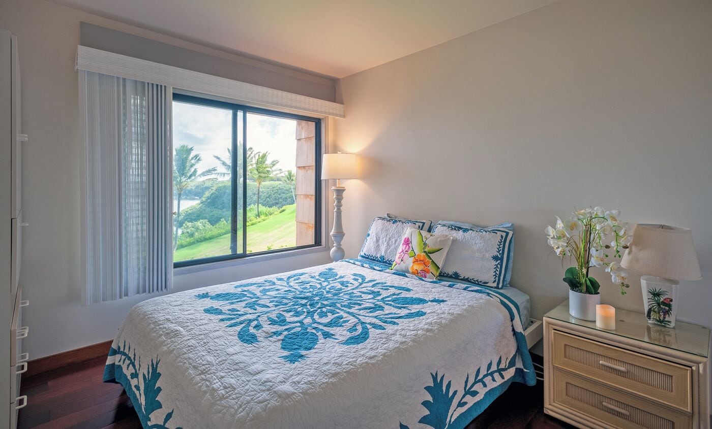 A bedroom with a large window, a bed with a blue and white quilt, a nightstand with a lamp, and a vase with flowers.