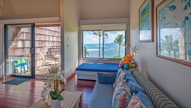 A room with a large window overlooking the ocean, a bed with blue bedding, a wicker sofa, and a small table with flowers. - Sealodge E6 (Princeville)