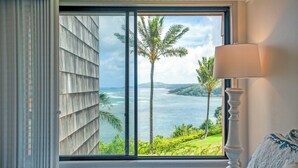 A room with a large window overlooking a coastal landscape with palm trees and a view of the sea. - Sealodge E6 (Princeville)