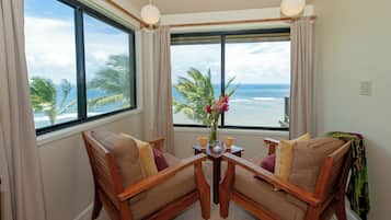 Quarto com duas cadeiras de madeira viradas para uma mesa, um vaso com flores e vista para o oceano através de janelas amplas.