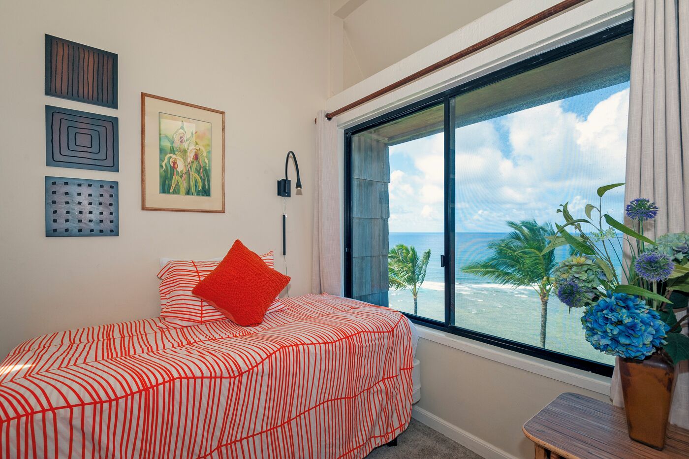 Ein Schlafzimmer mit einem großen Fenster, einem Bett mit gestreifter Bettwäsche, einem roten Kopfkissen und einer Vase mit blauen Blumen.