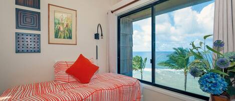 A bedroom with a large window, a bed with striped bedding, a red pillow, and a vase with blue flowers.