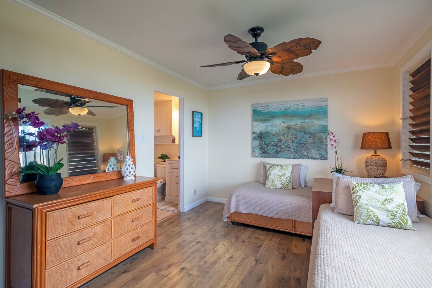 A bedroom with a wooden dresser, two beds, a ceiling fan, and a painting on the wall.